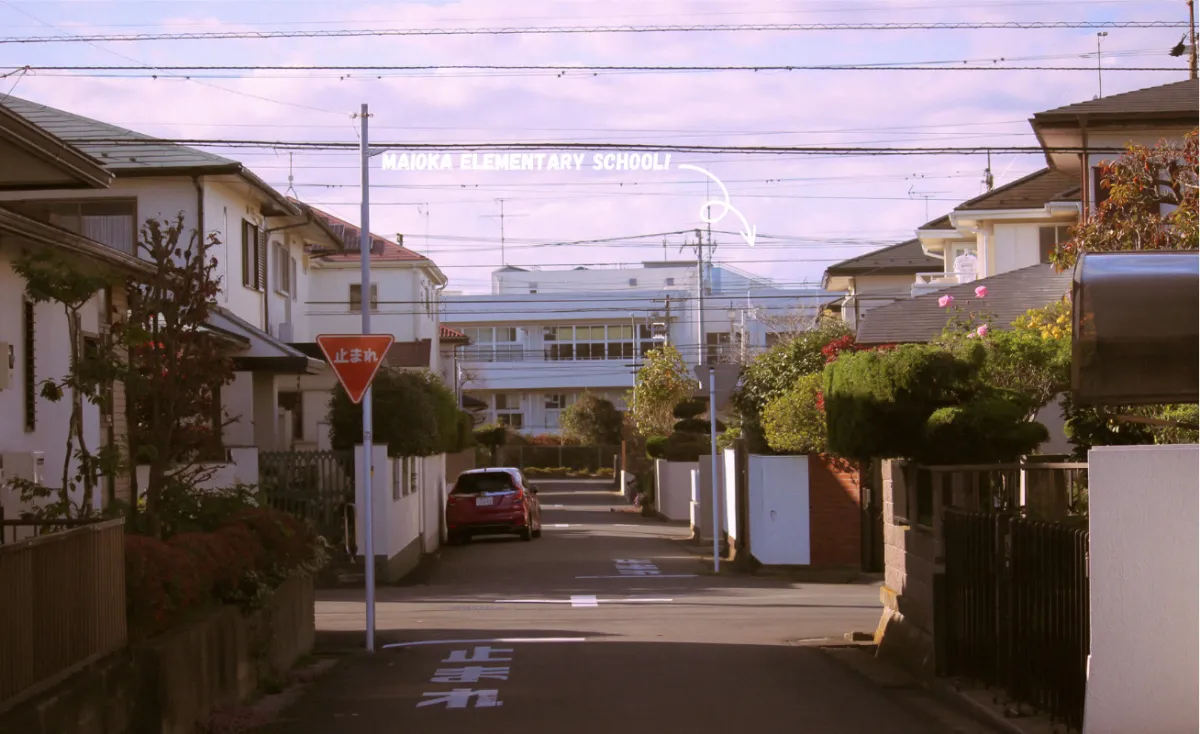 A quiet residential street in the Maioka area of Yokohama, Japan, under a wide sky, with a handwritten note reading MAIOKA ELEMENTARY SCHOOL! and an arrow pointing to the school at the end of the street.